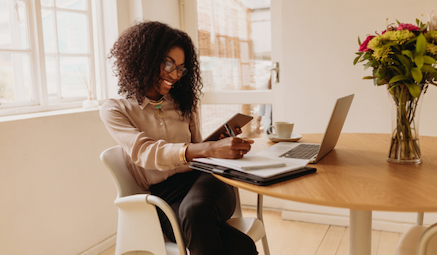 loan readiness program Hybrid Delivery A young Black woman wearing glasses and a business casual shirt, smiling and writing in a notebook while working on a laptop at a round table.